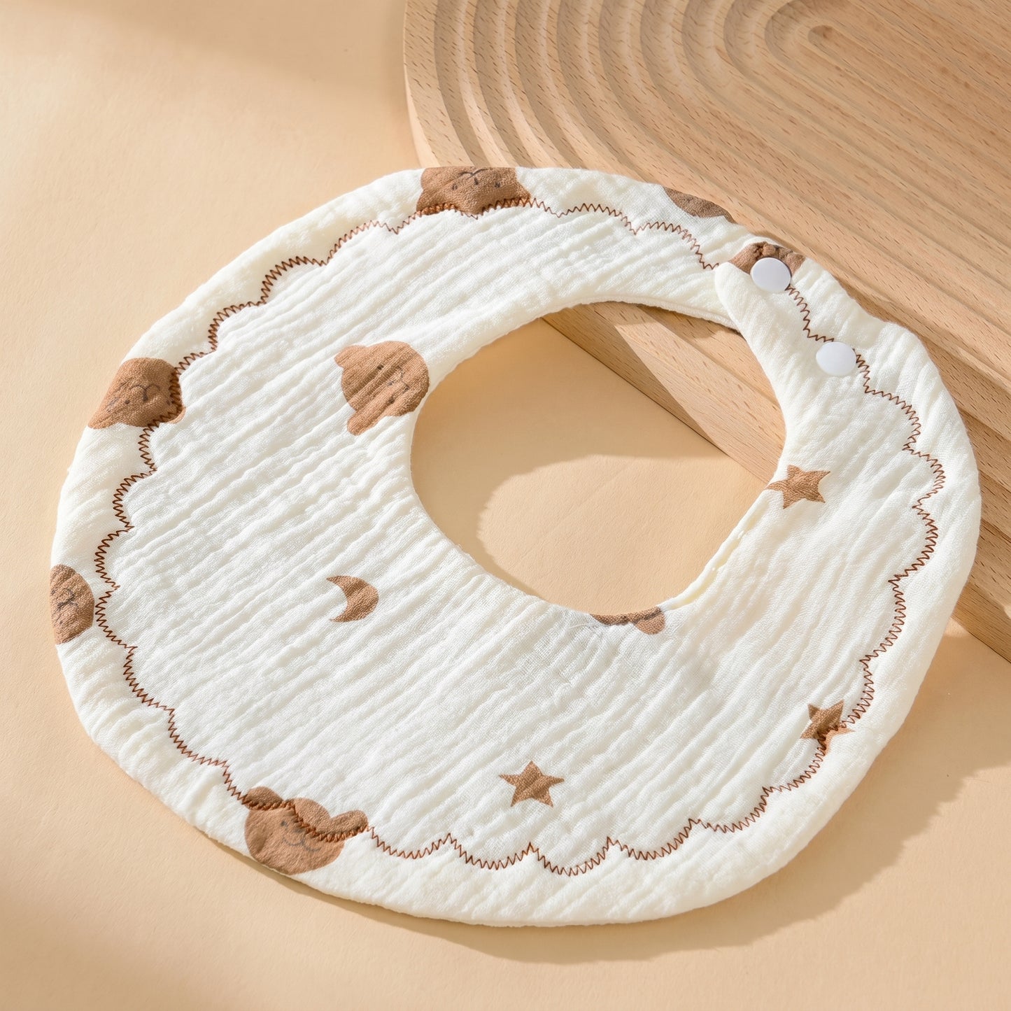 WhisperSoft™ cotton bib in cream with brown cloud and star patterns, featuring a scalloped edge and snap closures.