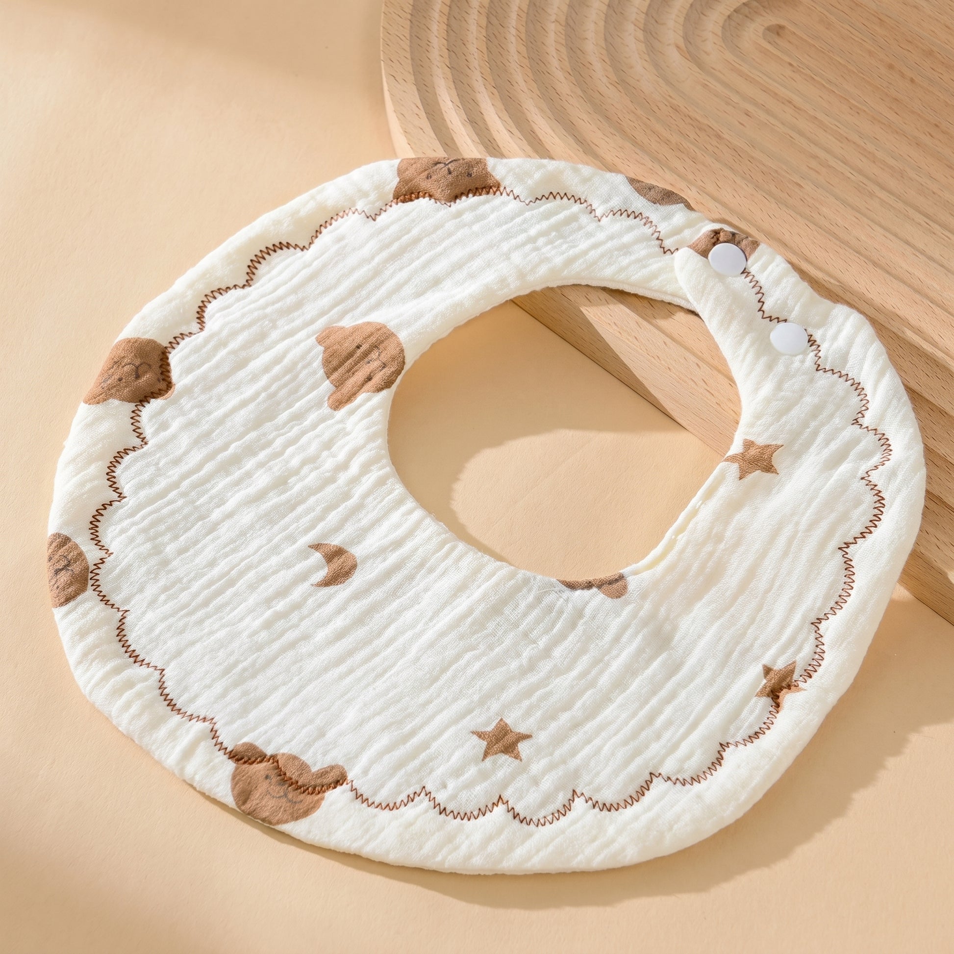 WhisperSoft™ cotton bib in cream with brown cloud and star patterns, featuring a scalloped edge and snap closures.