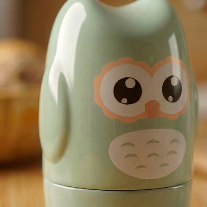 A close-up of a glossy light green owl-shaped container or decor piece with a cute printed owl face in peach, white, and dark brown, set against a warm blurred background.